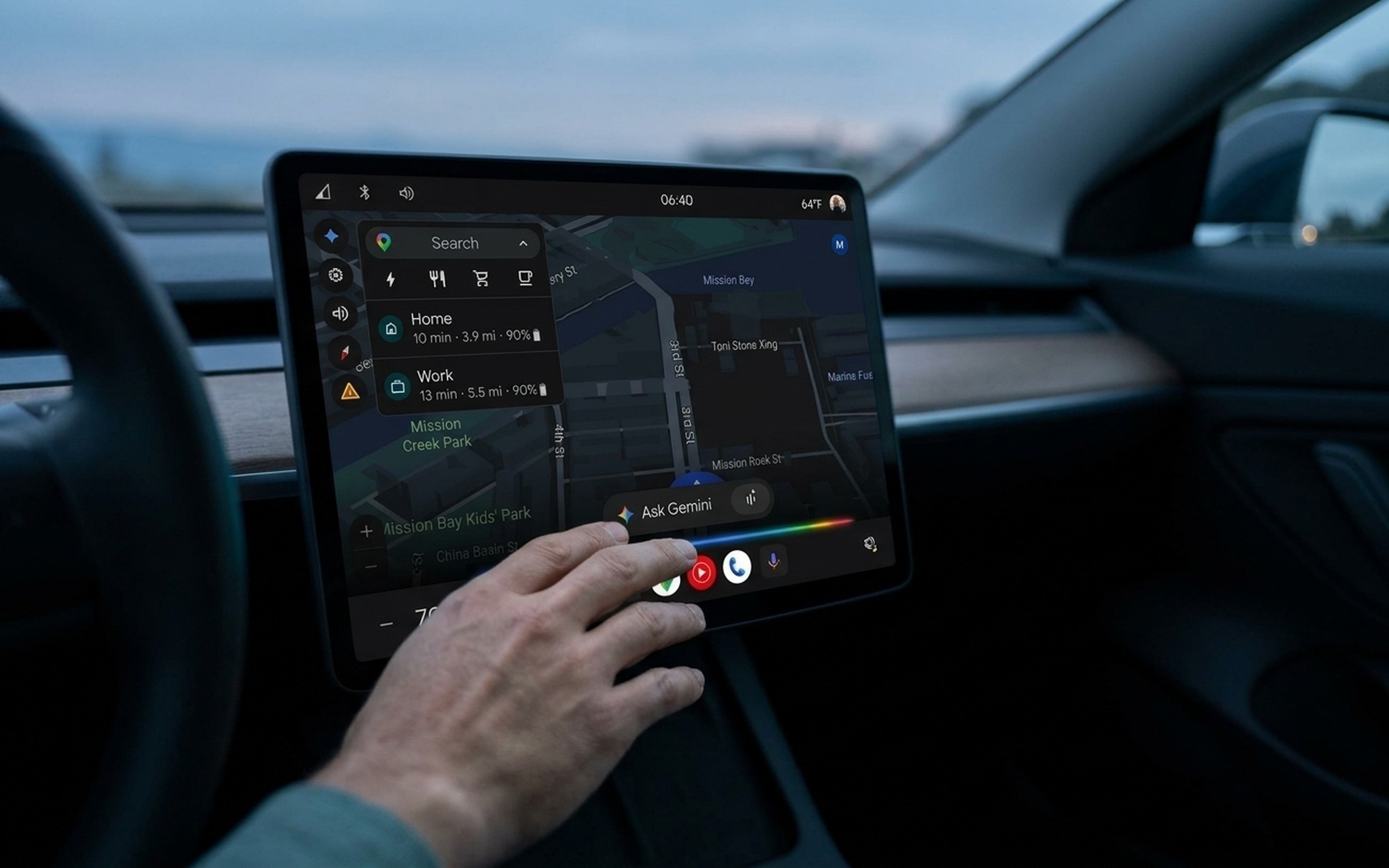 Driver's hand reaching toward car touchscreen showing Android Auto dark navigation map with Gemini AI Magic Cue voice assistant button