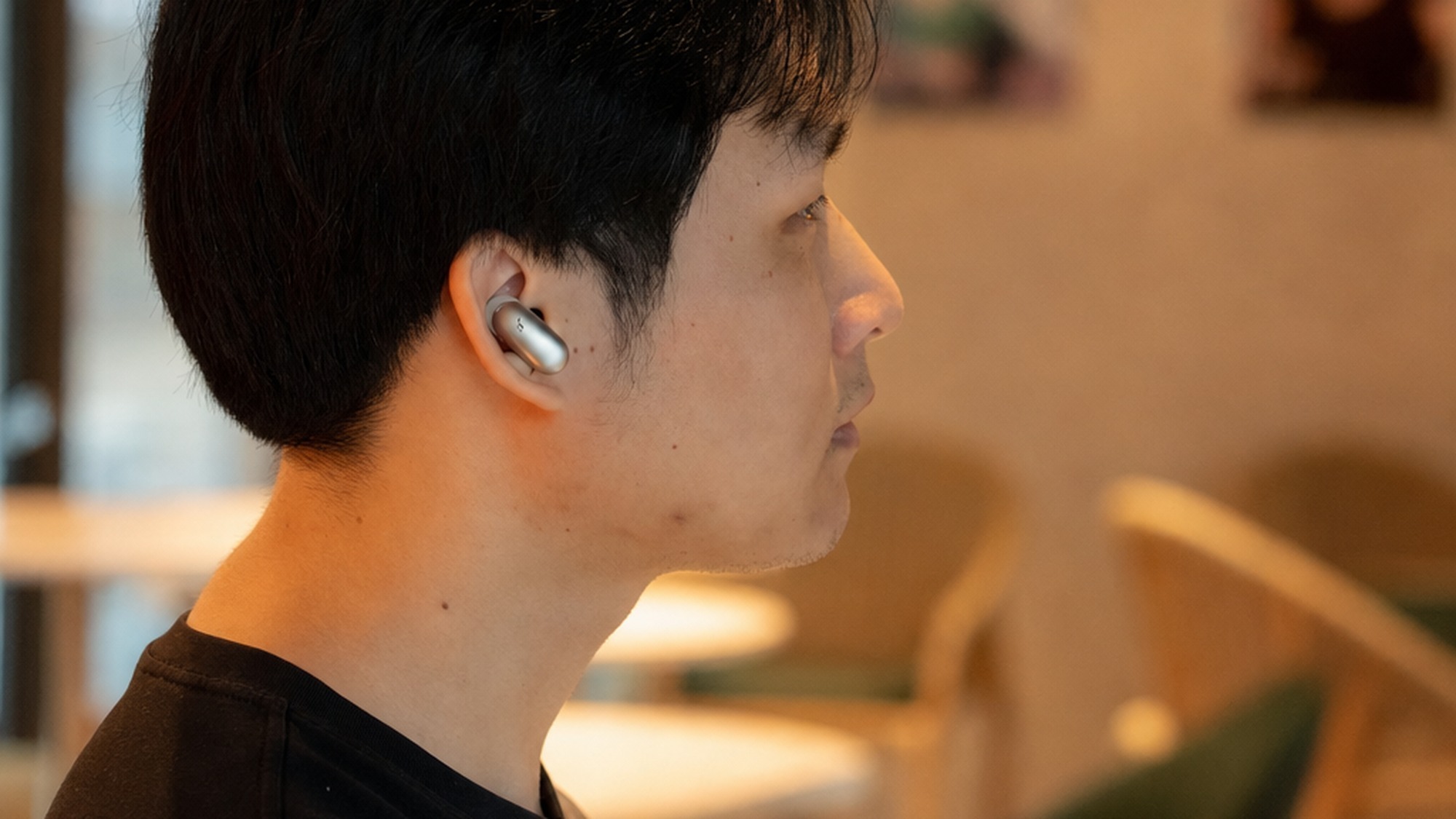 Side profile of a man wearing Anker Soundcore Liberty 5 Pro Max earbud in a warmly lit cafe interior with chairs