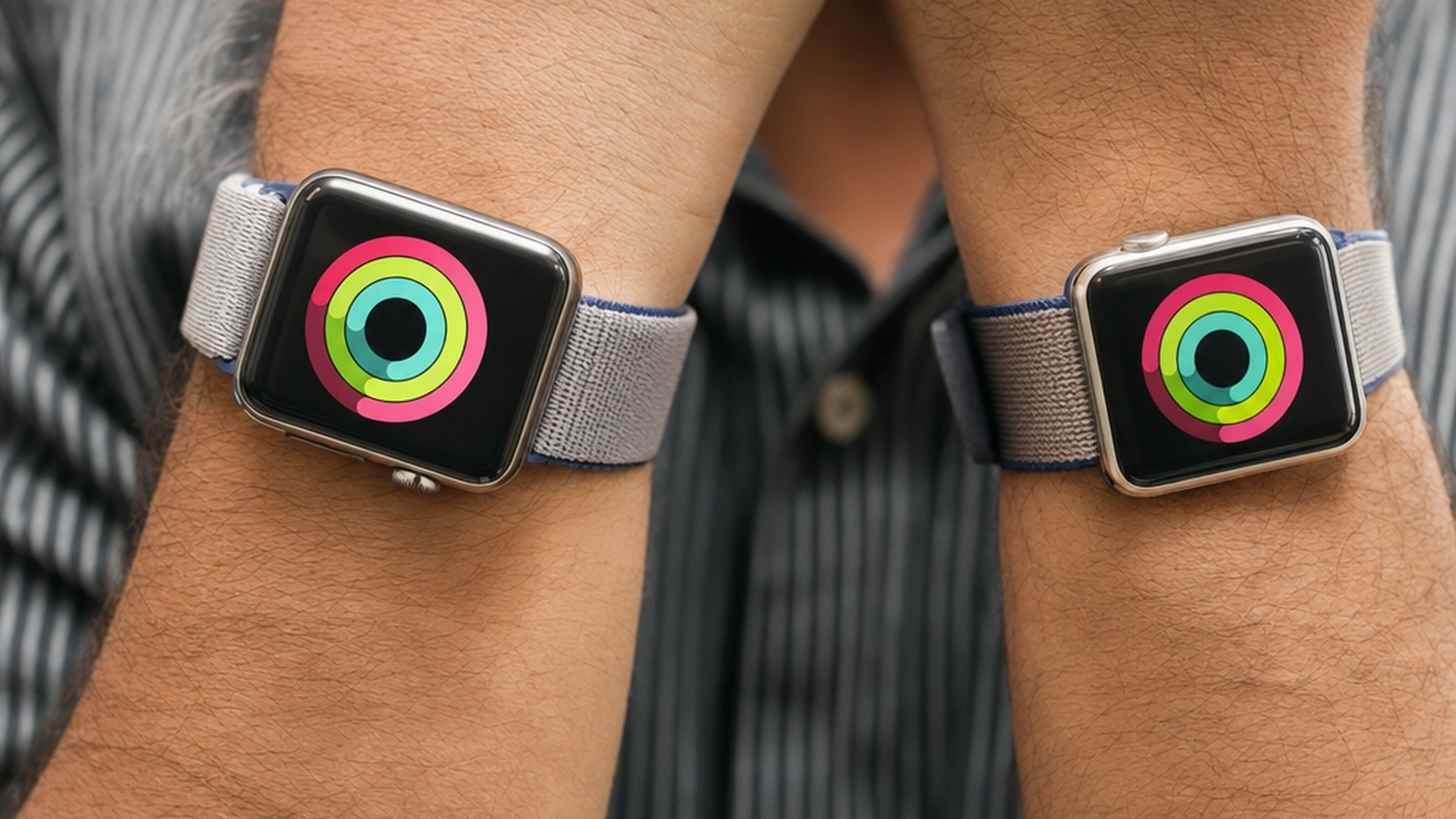 Two smartwatches with colorful activity ring graphics worn on two wrists, illustrating the Apple Watch companion device strategy.