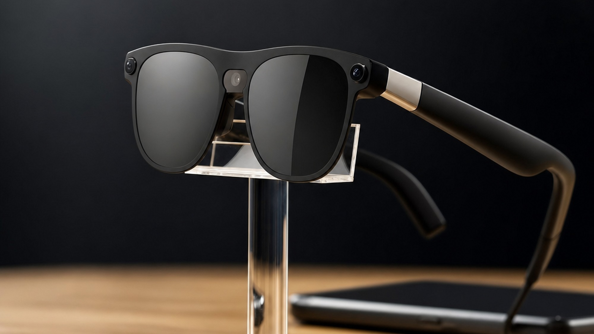Modern black acetate smart glasses with small temple camera modules displayed on a clear acrylic stand beside a blurred smartphone.