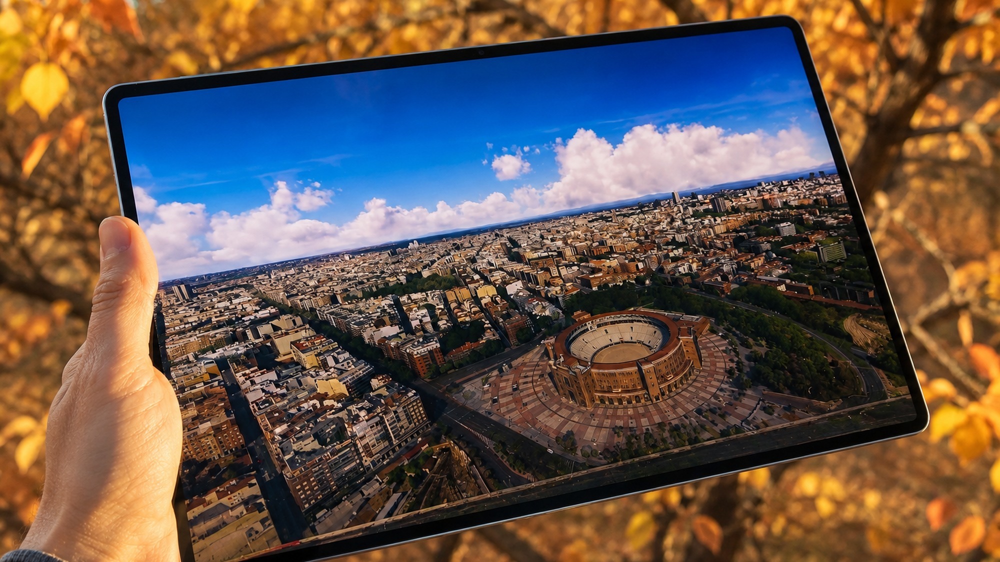 Hand holding the Huawei MatePad Pro Max outdoors, its 13.2 inch OLED display showing a bright aerial video against autumn branches.
